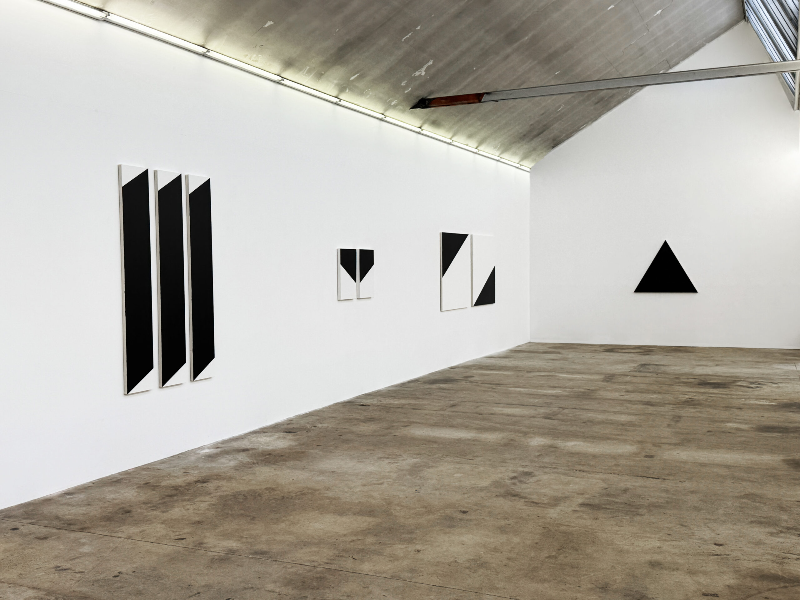 Jean-Charles Kien's geometric art exhibition view, featuring diverse black and white abstract paintings, including a set of three large vertical canvases and two smaller square works on a long white wall, alongside a solitary triangle composition, all displayed on concrete floors within a modern studio gallery, during La Regionale 2025
